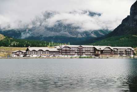 [A view across a body of water at a five-story mountain chalet type brown building with a light-color roof. Beside and behind the building are very tall mountains. Mists cover a portion of the mountain behind the building.]